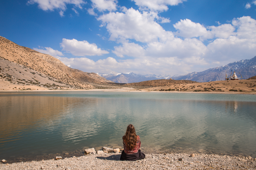 Woman sitting in frong of a lake with mountains in the back.